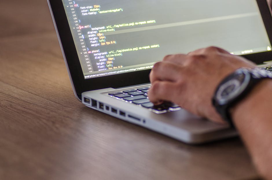 A close-up of a person’s hand typing on a silver laptop keyboard placed on a wooden desk, with the screen displaying lines of HTML or coding language. The individual is wearing a black wristwatch wi