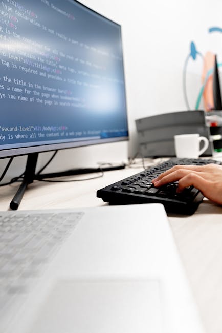 A close-up view of a workspace in a gardening company office, featuring a computer monitor displaying lines of coding or digital data, with a person's hand typing on a black keyboard. In the backgroun